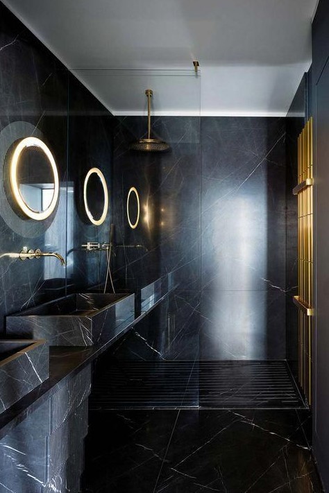 a minimalist black and gold bathroom clad with black marble, with lit up mirrors, gold accessories and fixtures
