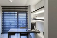 a minimalist home office with an open shelving unit with lights, a built-in desk and a black chair plus a floor to ceiling window