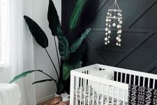 a modern boho nursery with a black accent wall, a white crib, printed textiles, layered rugs, a white chair and a woven pendant lamp