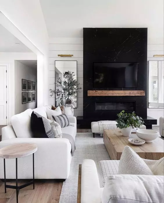 a modern farmhouse living room with white seating furniture, a wooden coffee table, a black fireplace with a wooden mantel and greenery