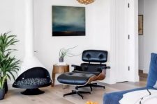 a modern living room with a blue low sofa, a black leather chair with a footrest, a white Malm fireplace and a wooden pendant lamp plus a pouf