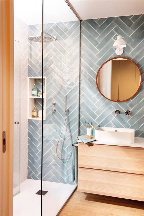 a modern neutral bathroom with blue chevron tiles, a light-stained vanity and a floor, a round mirror, a shower with a skylight