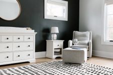 a monochromatic nursery with a black accent wall, a white dresser, a white chair and  a footrest, a nightstand and a cool printed rug