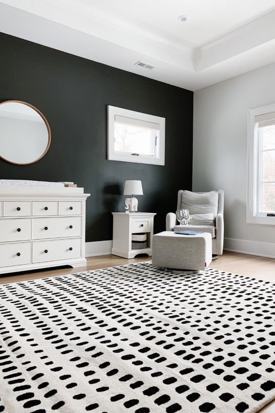 a monochromatic nursery with a black accent wall, a white dresser, a white chair and  a footrest, a nightstand and a cool printed rug