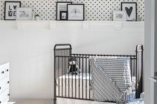 a monochromatic nursery with white paneled and polka dot walls, a black crib, a grey chair and a black pouf, a white dresser