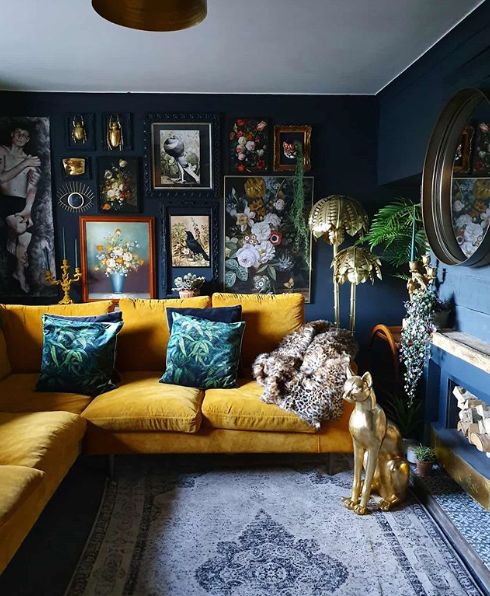 a moody and refined living room with navy walls, a bold yellow sofa, a fireplace with firewood and a round mirror plus gold decor