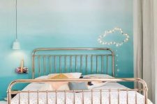 a pretty bedroom with an ombre turquoise wall, a metal bed with printed bedding, a printed rug, a cloud of string lights and pendant lamps