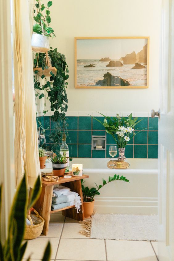 a pretty eclectic bathroom with green tiles, a neutral tub, a wooden stool, potted plants, a lovely artwork and neutral textiles