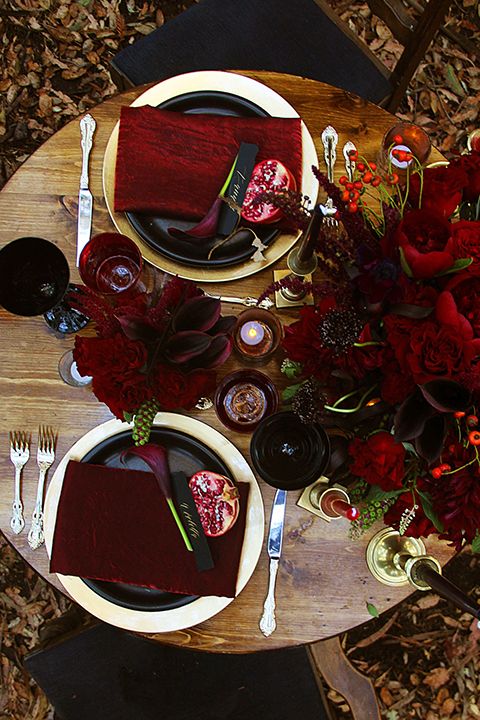 a refined deep red, black and gold Halloween tablescape with deep-toned blooms, black plates and glasses, greenery and gold candleholders
