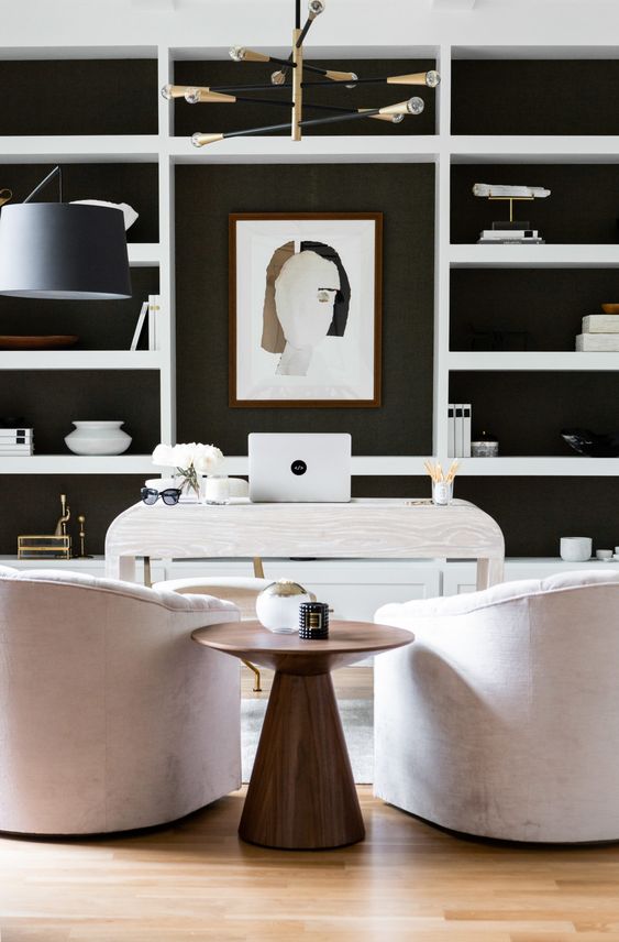 a refined modern home office with a black accent wall and white shelves on it, a white stone desk and neutral chairs, a side table and a lovely chandelier