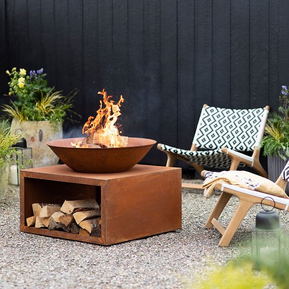 a simple and stylish outdoor fire pit space with light stained chairs, a metal bow and a fire bowl on top plus potted blooms