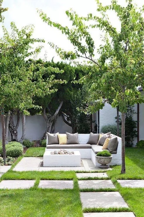 a small and lovely neutral outdoor space with a corner bench with neutral pillows, a box shaped fire bowl with rocks is cool