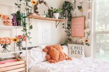 a small boho teen bedroom with a grey bed and white bedding, a shelving unit and an open shelf, lots of potted plants and blooms