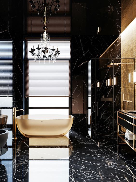 a sophisticated black and gold bathroom with black marble, a gold accent wall, a gold lucite bathtub, a black chandelier and built-in lights