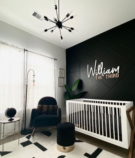 a stylish contrasting nursery with a black accent wall, a white crib with black and white bedding, a black chair and a pouf, a potted plant and a chandelier