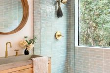 a welcoming modern bathroom with mint skinny tiles, a light-stained vanity, a round mirror and a window in the shower space