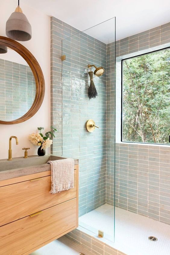 a welcoming modern bathroom with mint skinny tiles, a light stained vanity, a round mirror and a window in the shower space