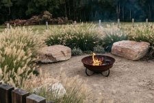 a wild modern outdoor space with greenery and grasses, large rocks as seats and a metal fire bowl on a stand