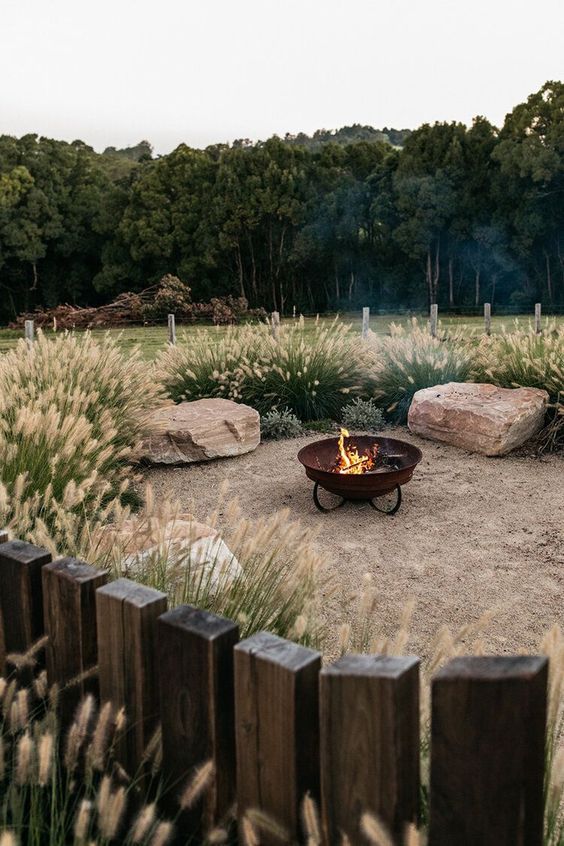 a wild modern outdoor space with greenery and grasses, large rocks as seats and a metal fire bowl on a stand