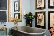 an eclectic bathroom with light blue walls, a large mirror in a gilded frame, a grid gallery wall, a grey stone tub and a bold floor and a rug