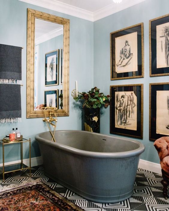 an eclectic bathroom with light blue walls, a large mirror in a gilded frame, a grid gallery wall, a grey stone tub and a bold floor and a rug