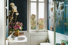 an eclectic bathroom with navy walls and a gallery wall of jewelry, a vintage free-standing sink, glam lamps and gold accessories