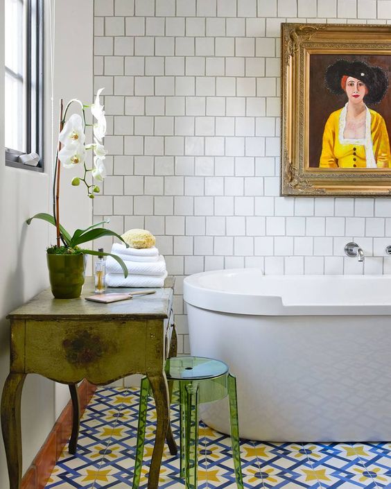 an eclectic bathroom with square and colorful tiles, an oval tub, a vintage vanity, a green acrylic stool and an artwork