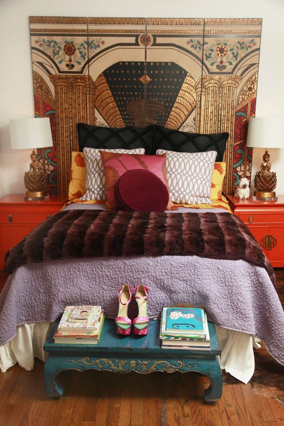 an eclectic bedroom with a bed with colorful bedding, red nightstands, a blue bench, a bold artwork, catchy Asian style lamps