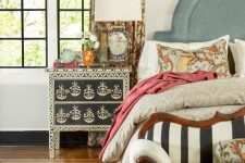 an eclectic bedroom with a blue upholstered bed, colorful bedding, a stenciled nightstand, a striped loveseat, printed curtrains