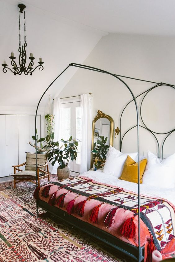 an eclectic bedroom with a large forged bed with colorful boho bedding, a bold boho rug, potted plant, a vintage chandelier on chain