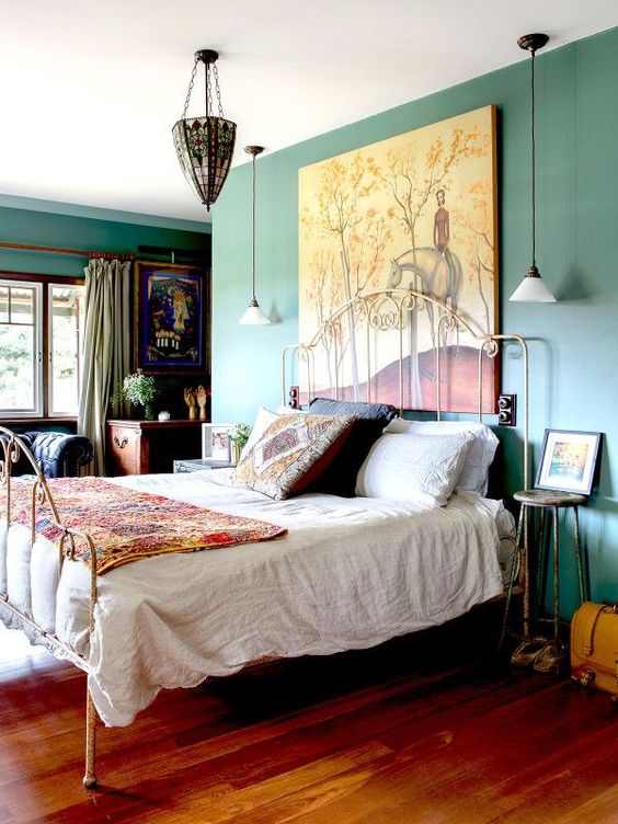 an eclectic bedroom with green walls, a forged bed with colorful and printed bedding, forged stools, a dark-stained desk, a chic chandelier and an oversized artwork