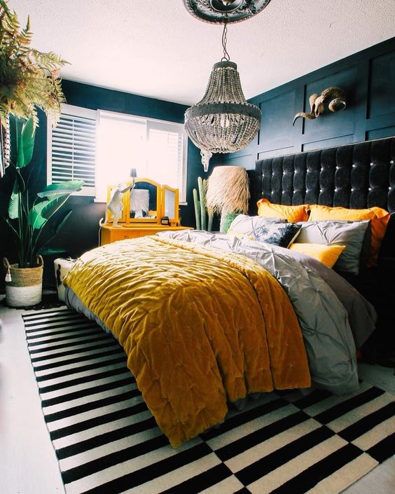 an eclectic bedroom with navy paneled walls, a black upholstered bed, colorful yellow bedding, a yellow vanity, potted statement plants and a beaded chandelier