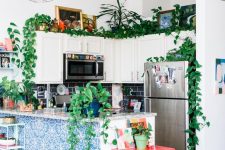 an eclectic kitchen with white cabinets, a navy tile backsplash, a kitchen island with blue printed tiles, potted plants and bright artwork