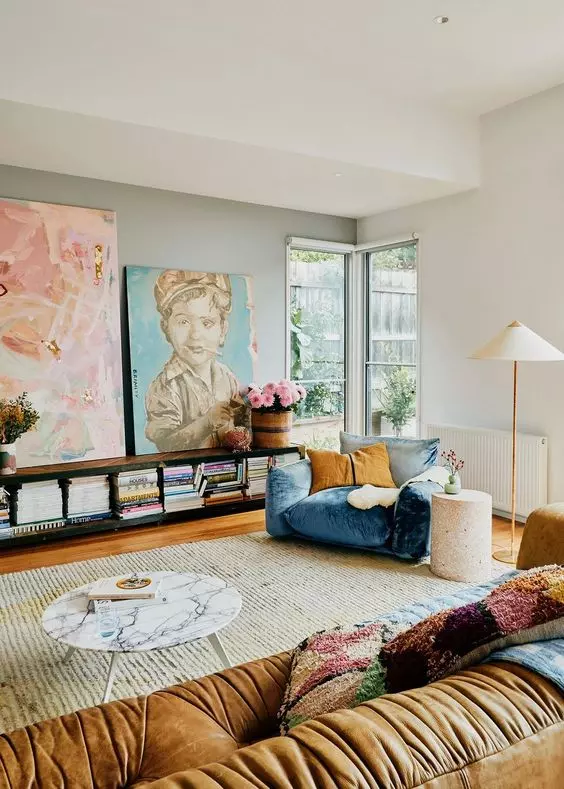 an eclectic living room with a blue and mustard chair, a beige sofa, colorful pillows, a bookshelf, a marble table and artwork