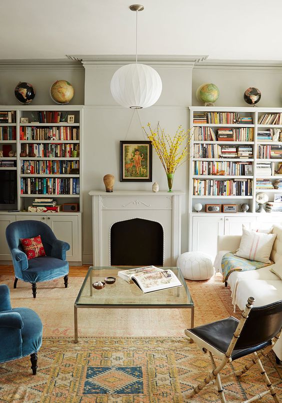 an eclectic living room with built-in bookshelves, a fireplace, a creamy sofa, blue chairs, a black one, a glass coffee table and printed rugs