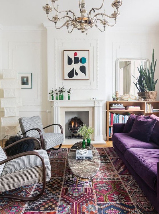 an eclectic living space with a purple sofa, printed rockers, a faux fireplace, a large boho rug and a vintage chandelier