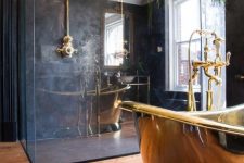 an elegant bathroom with black walls, a glass enclosed shower space, a polished gold bathtub, gold fixtures and greenery