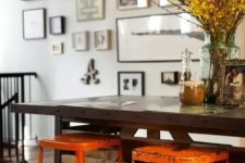 an industrial dining space with a dark table, orange metal stools and a gallery wall, blooming branches in a vase