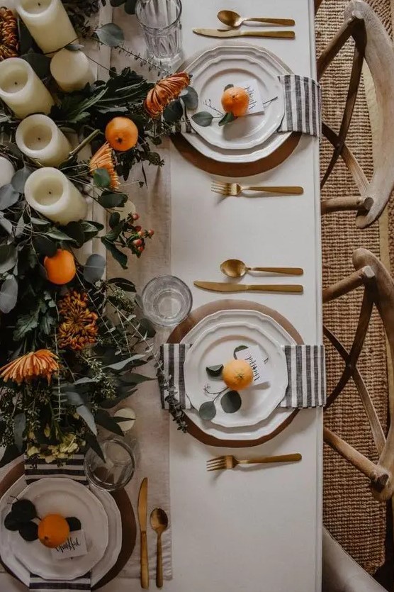 a beautiful Thanksgiving tablescape with striped napkins, gold cutlery, a greenery runner, blooms, citrus and candles