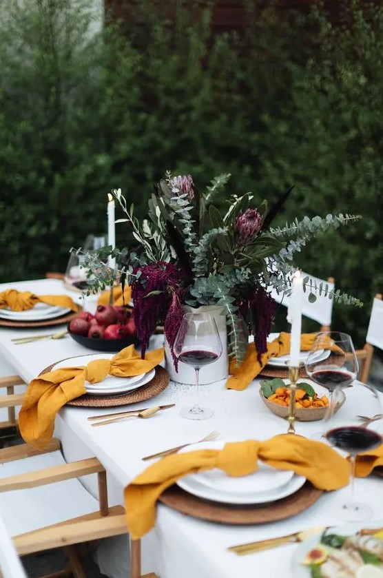 a bright modern Thanksgiving table with greenery and deep purple blooms, yellow napkins, woven chargers and gold cutlery rocks