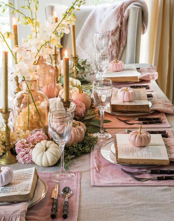 a chic glam Thanksgiving table setting with pink placemats, vintage books, pastel pumpkins and blooms, tall candles