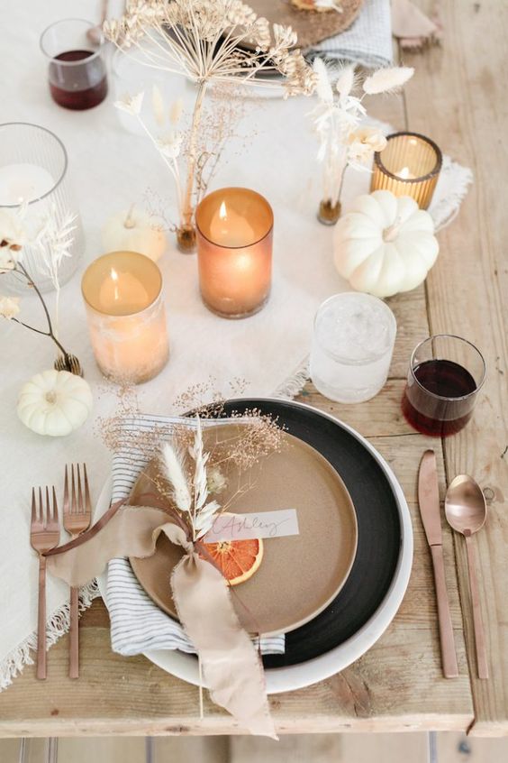 a catchy modern meets boho Thanksgiving tablescape with a neutral runner, black and beige plates, copper cutlery and candles, grasses and white pumpkins