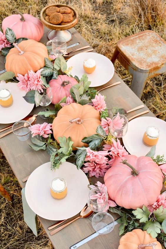 a chic modern Thanksgiving table setting with orange and pink pumpkins, pink blooms and greenery, neutral plates and green napkins