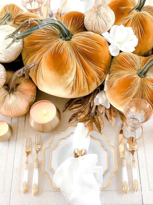 a glam Thanksgiving tablescape with orange and rust velvet pumpkins, gilded leaves, white and gold plates and refined cutlery