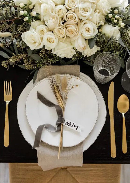 a gorgeous glam Thanksgiving table setting with a black uncovered table, white plates, gold cutlery, white florals and greenery and wheat