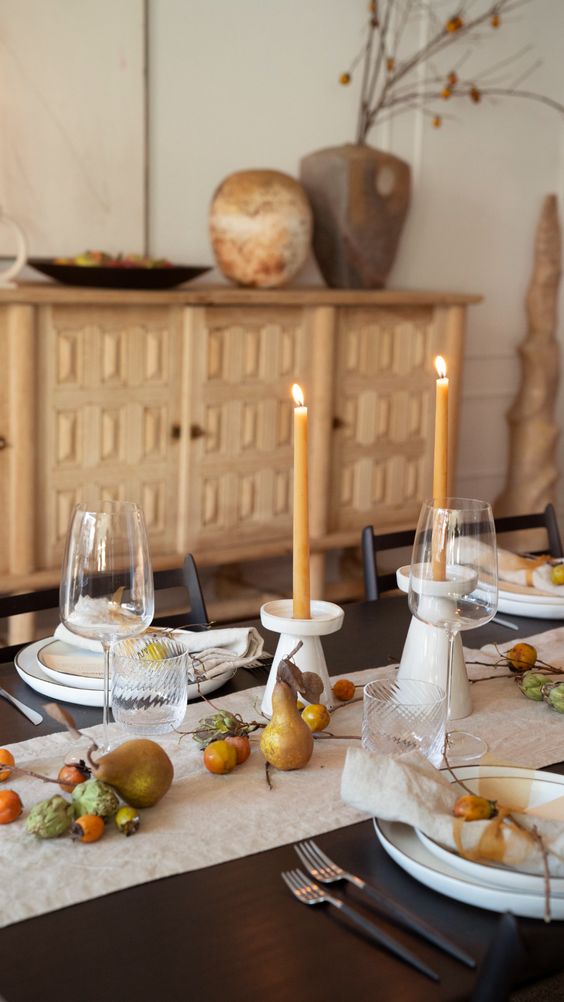a modern organic Thanksgiving table setting with a neutral runner and napkins, neutral plates, black cutlery, yellow candles and fruit right on the table