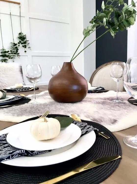 a modern Thanksgiving tablescape with a faux fur runner, black woven placemats, white porcelain and pumpkins, a brown vase with greenery