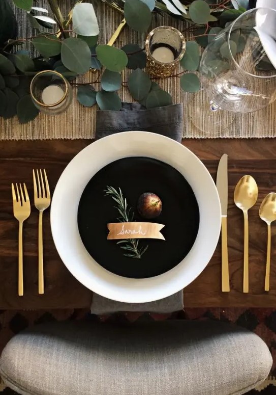 a modern Thanksgiving tablescape with a woven table runner, greenery, gold glitter, gold cutlery and black and white plates