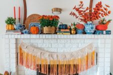 a bright fall mantel with bold blooms, potted greenery, orange, grey and neutral pumpkins, colorful fringe and some bold books