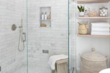 09 a neutral farmhouse bathroom with floating shelves, a basket with a lid, a shower space enclosed in glass and a wooden stool